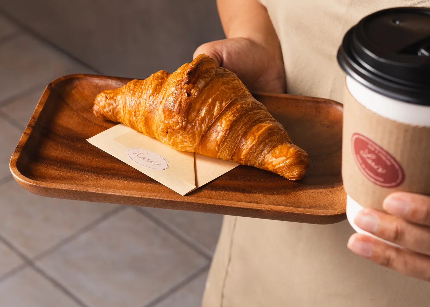 Croissant sobre bandeja de madera y café para llevar, en manos de la casa